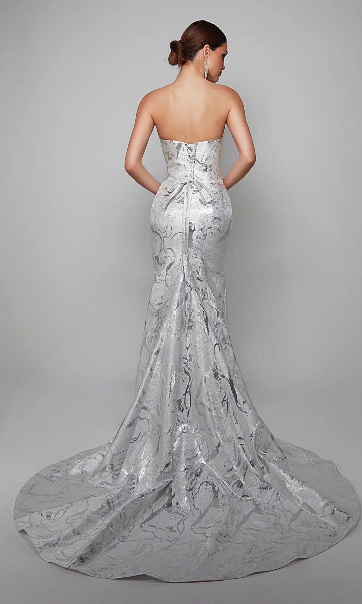 A woman faces away wearing the Alyce 7055, a long white strapless gown with a fitted bodice and flowing train—ideal for a mother-of-the-bride look against a plain backdrop.